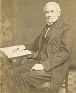 Stained photo of seated white man in three piece suit, high collar and bow tie, his right hand resting on an open book.